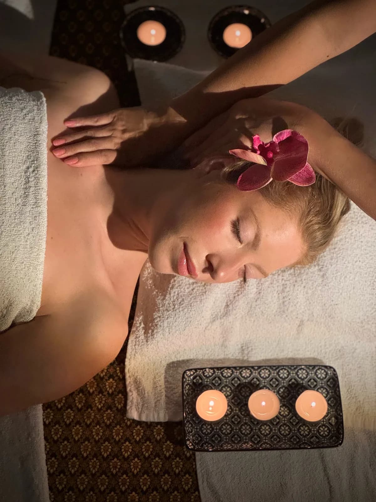 Cliente rilassata con gli occhi chiusi e un'orchidea tra i capelli mentre la terapista preme sulla spalla durante un Massaggio Aromatico, circondata da candele